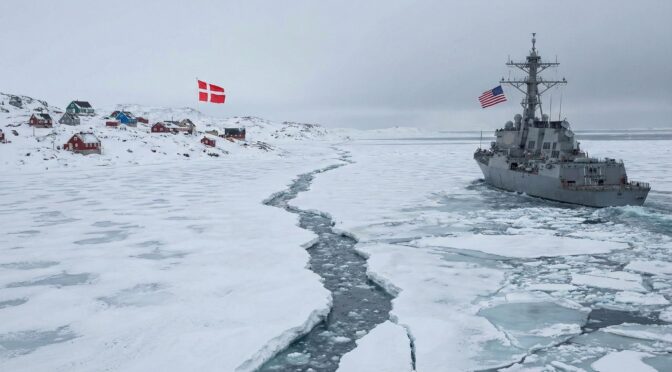 US Navy ship sailing through icy waters near Greenland, with Danish flag visible on the shore.