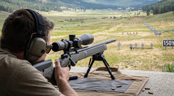 Man aims a rifle at a target range with hills in the background.