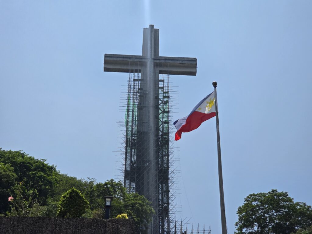 Philippine flag waves near the shrine's massive cross under construction with scaffolding.