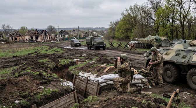 Soldiers in trench with military vehicles and damaged buildings in background.