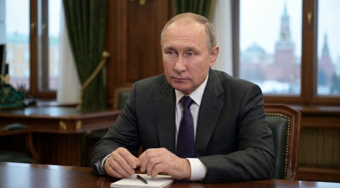 Vladimir Putin in a dark suit and tie sits at a desk with a notebook and pen.