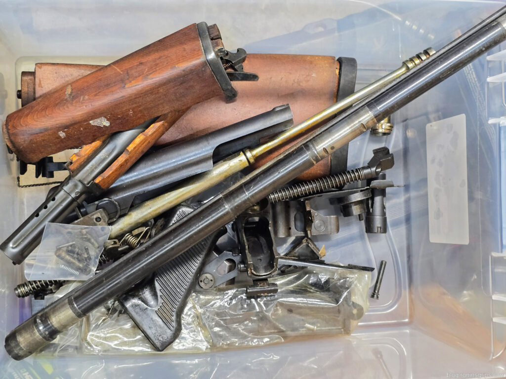 Disassembled wooden rifle stock and metal gun parts in a clear plastic bin.