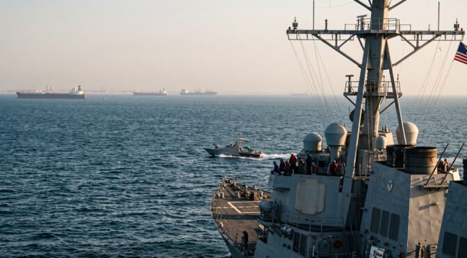 US Navy destroyer sails alongside a patrol boat in the ocean, with cargo ships in the distance.